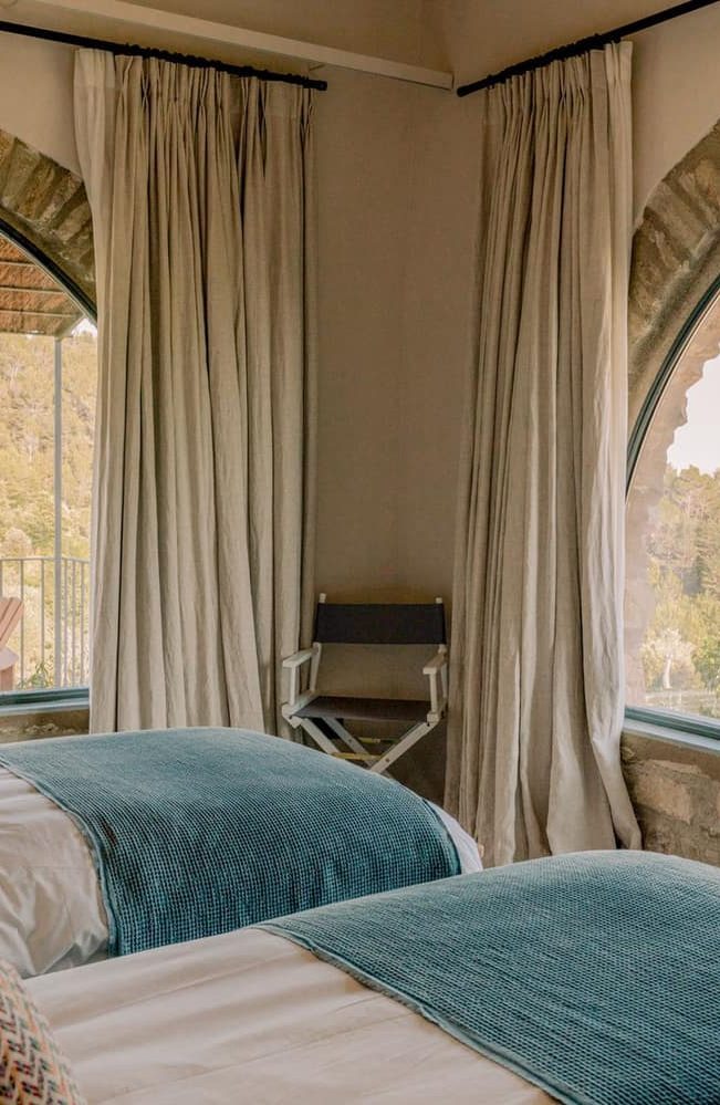 A twin bedroom with teal throws and tall linen curtains by arched windows