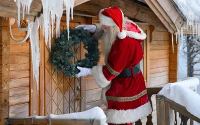 Santa hanging a Christmas wreath on the door of a snow-covered log cabin hung with icicles