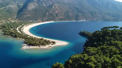 The Blue Lagoon at Ölüdeniz from the mountain above — a curving sand spit between a turquoise lagoon and the deep blue sea, pine-covered mountains behind