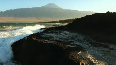 Black volcanic rock on the Tenerife coast, with Atlantic surf breaking white over it and the peak of Teide hazy in the distance.